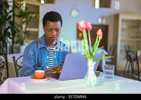 Donna nera con i capelli corti che indossa una giacca in denim che lavora su un computer portatile mentre è seduta al tavolo del ristorante Foto Stock