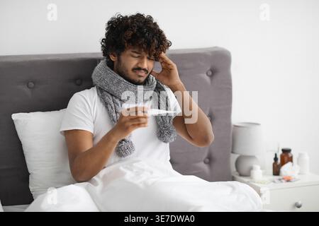 Uomo asiatico malato con capelli ricci che indossa sciarpa intorno al collo toccando la testa e tenendo il termometro che controlla la temperatura corporea mentre si siede sul letto io... Foto Stock