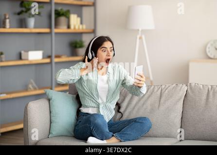 Donna indiana scioccata con heafoni che usa lo smartphone seduto con le gambe incrociate sul divano nel soggiorno di casa Foto Stock