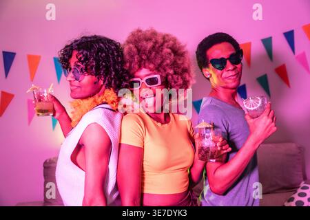 Gruppi diversi di amici con occhiali da sole che tengono in mano occhiali da cocktail che ballano insieme nella stanza di luce rosa di casa Foto Stock