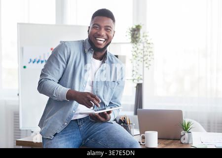 Allegro uomo d'affari nero che usa un tablet seduto su un tavolo con un computer portatile in ufficio Foto Stock
