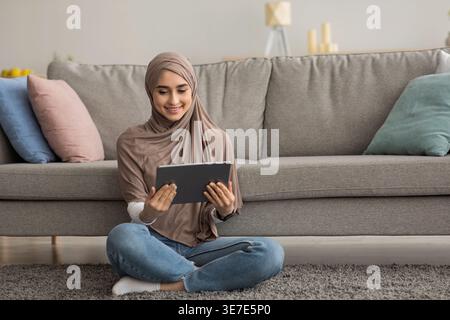 Donna araba che indossa un hijab con un tablet appoggiato sul divano seduto con le gambe incrociate sul tappeto nel soggiorno di casa Foto Stock