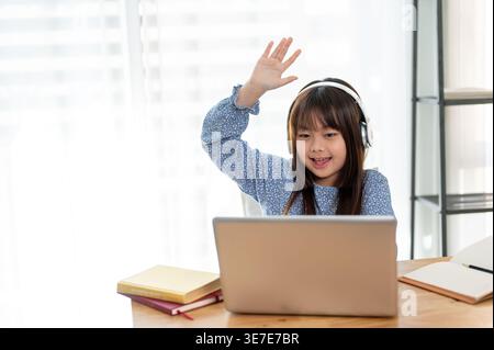 Ragazza asiatica che indossa le cuffie alzando la mano mentre si tiene una lezione online seduta a tavola con i libri a casa Foto Stock