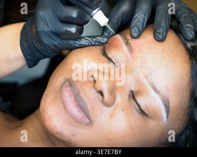 Mani di una cosmetologa ispanica che indossa guanti usando un pennello per definire la forma delle sopracciglia della cliente femminile nera in salone Foto Stock