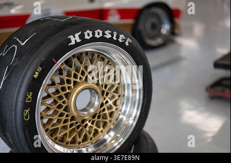 Primo piano di un pneumatico Hoosier Racing slick montato su un cerchio in lega con mesh dorata e mozzo con blocco centrale, caratterizzato da un'auto da corsa sfocata sullo sfondo di una Foto Stock