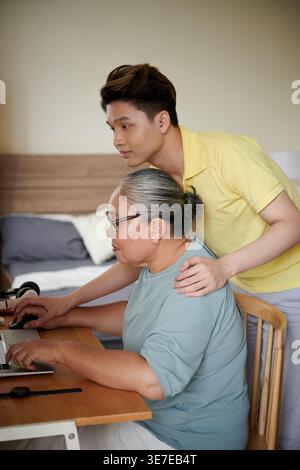 Uomo asiatico in piedi e inclinato in avanti dietro la nonna seduto al tavolo insegnandole come usare il laptop a casa Foto Stock