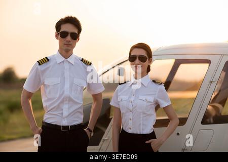 Piloti cinesi maschili e femminili con occhiali da sole in piedi vicino all'elicottero durante il tramonto Foto Stock