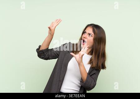 Donna ispanica spaventata che indossa un blazer che si muove e guarda in alto mentre si trova sullo sfondo verde chiaro in studio Foto Stock
