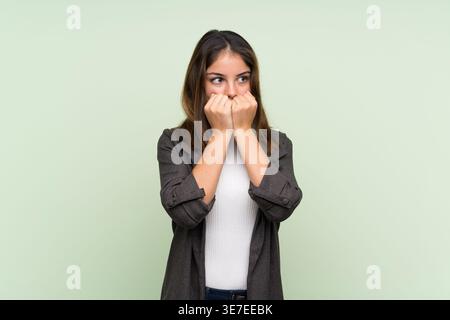 Donna ispanica che indossa un blazer che guarda lontano e mette le mani alla bocca mentre sta in piedi su uno sfondo verde chiaro in studio Foto Stock