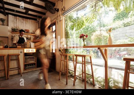 Barista asiatico che lavora al bancone mentre un cliente donna cammina nel bar Foto Stock