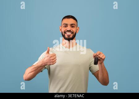 Sorridente uomo mediorientale con barba che tiene la chiave dell'auto e mostra i pollici in piedi su sfondo blu in studio Foto Stock