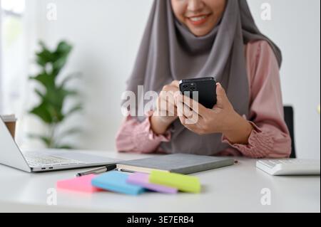 Donna asiatica sorridente in foulard con smLifestylephone seduto vicino al laptop al tavolo in ufficio Foto Stock