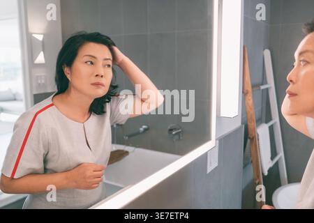 Perdita di capelli donna asiatica matura toccando i capelli o colorando i capelli grigi guardando se stessa nello specchio di casa. Beautifiul cinese senior lady in... Foto Stock