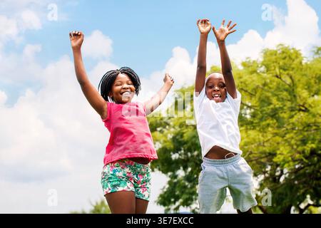 Felice ragazzo e ragazza neri che saltano con le mani in alto nel parco nella giornata di sole Foto Stock