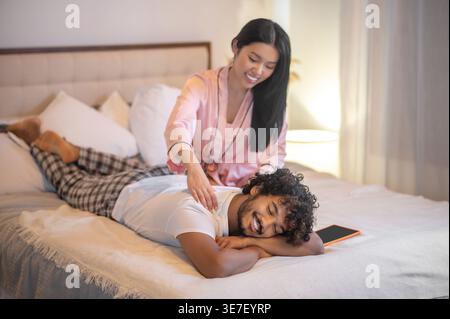 Donna ispanica sorridente che fa un massaggio alla schiena al fidanzato sdraiato sul letto Foto Stock
