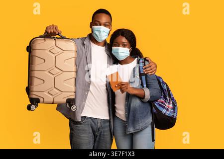 Coppia nera che indossa maschere facciali che trasportano zaino e valigia in piedi su sfondo giallo in studio Foto Stock