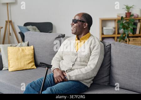 Happy Black cieco che indossa occhiali da sole seduto sul divano di casa Foto Stock