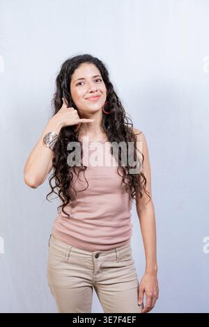 Donna ispanica che fa gesti telefonici con le dita in piedi davanti allo sfondo bianco in studio Foto Stock