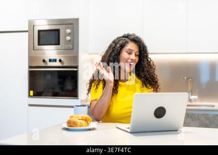 Donna ispanica con capelli ricci che partecipa a una riunione online utilizzando un computer portatile mentre fa colazione al bancone della cucina a casa Foto Stock