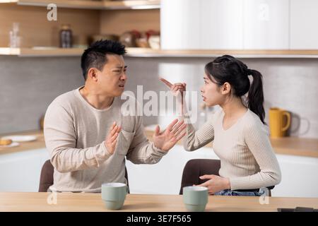 Donna asiatica arrabbiata che litiga con il marito gridando l'un l'altro e gestendo mentre si siede a tavola e beve un caffè in cucina a casa Foto Stock