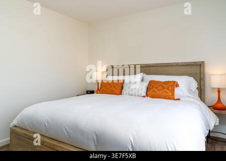 Moderna camera da letto bianca con telaio in legno, testiera, biancheria da letto, cuscini decorativi con accenti arancioni, lampade Foto Stock