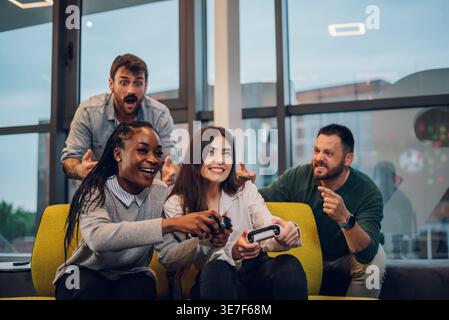 Donne nere e caucasiche sedute sul divano tenendo le console che giocano a videogiochi con colleghi maschi che tifanno dietro di loro in ufficio Foto Stock