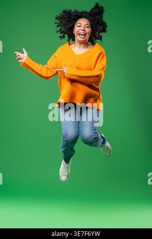 Donna nera con le dita ricciolate lateralmente mentre salta sullo sfondo verde in studio Foto Stock