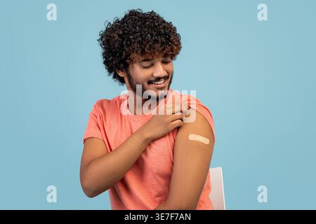 Uomo indiano barbuto con capelli ricci che mostra il braccio vaccinato mentre si siede sulla sedia di fronte allo sfondo blu in studio Foto Stock