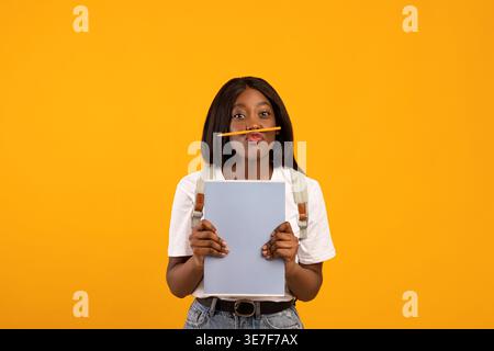 Donna nera con zaino che bilancia la matita sotto il naso che tiene libri in piedi su sfondo giallo in studio Foto Stock