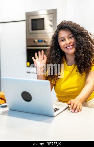 Donna ispanica con capelli ricci che partecipa a una riunione online utilizzando un computer portatile mentre fa colazione al bancone della cucina a casa Foto Stock