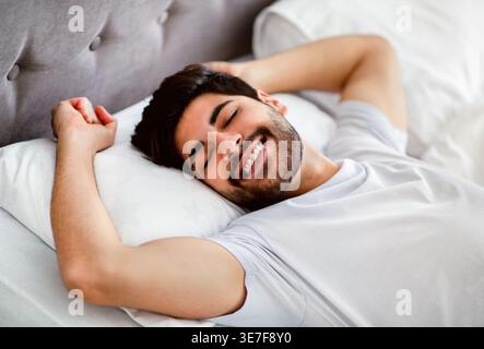 Uomo arabo sorridente con la barba che indossa una camicia bianca che allunga le braccia con gli occhi chiusi mentre è sdraiato sul letto in camera da letto di casa Foto Stock