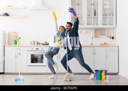 Uomo mediorientale e donna musulmana in hijab che si diverte a tenere il mocio e le scopa cantando mentre si sta in piedi e pulisce il pavimento della cucina a casa Foto Stock
