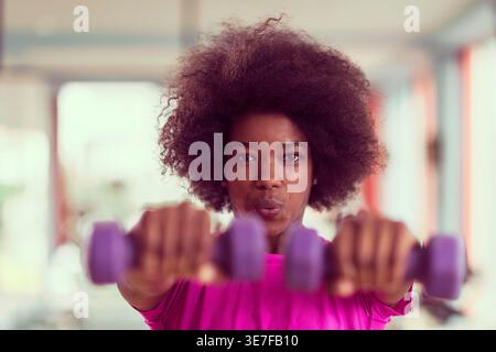 Donna nera con afro che fa esercizio fisico usando i manubri in piedi in palestra Foto Stock