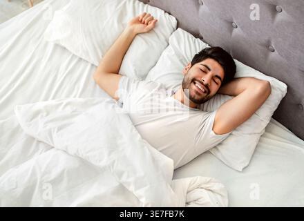 Uomo arabo sorridente con la barba che indossa una camicia bianca che allunga le braccia con gli occhi chiusi mentre è sdraiato sul letto in camera da letto di casa Foto Stock
