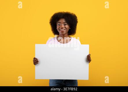 Femmina nera con afro che tiene in mano un cartone bianco su sfondo giallo in studio Foto Stock
