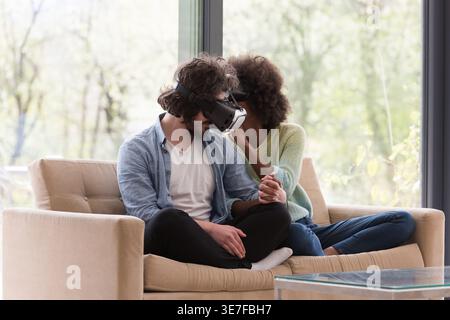 Coppia nera e caucasica che indossa occhiali VR che si tengono per mano seduti insieme sul divano e sperimentano la realtà virtuale in salotto con gl... Foto Stock