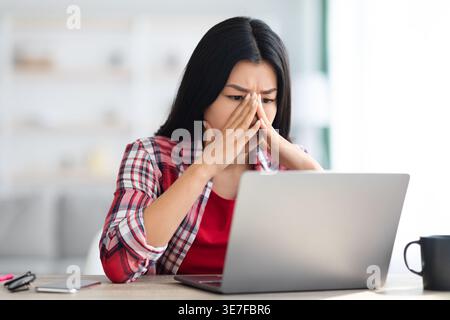 Donna asiatica stressata con le mani sul viso che guarda il laptop seduto a un tavolo a casa Foto Stock