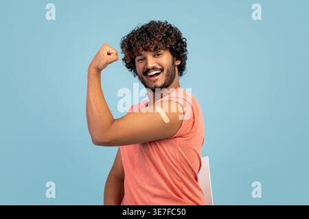 Uomo indiano barbuto con capelli ricci che mostra il braccio vaccinato mentre si siede sulla sedia di fronte allo sfondo blu in studio Foto Stock