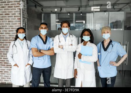 Gruppo diversificato di medici che indossano maschere mediche in piedi insieme davanti alla porta di vetro in ospedale Foto Stock