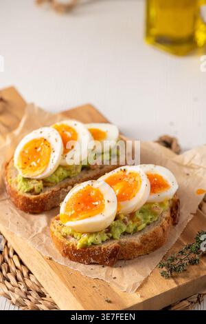 Due fette di lievito madre croccante ricoperte di purè di avocado e uova sode soffici perfettamente marmellate. Scatto ad angolo alto con luce naturale calda e ru Foto Stock