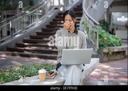 Una donna asiatica stanca si siede all'aperto, sentendosi esausta e sovraccarica di lavoro durante il lavoro sul suo laptop. Foto Stock