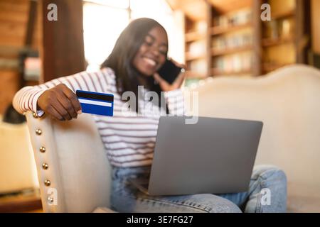 Adolescente di colore con carta di credito che riceve una telefonata seduto con il computer portatile sul divano nel soggiorno di casa Foto Stock