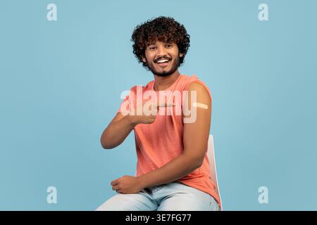 Uomo indiano barbuto con capelli ricci che punta il dito al braccio vaccinato mentre si siede sulla sedia di fronte allo sfondo blu in studio Foto Stock