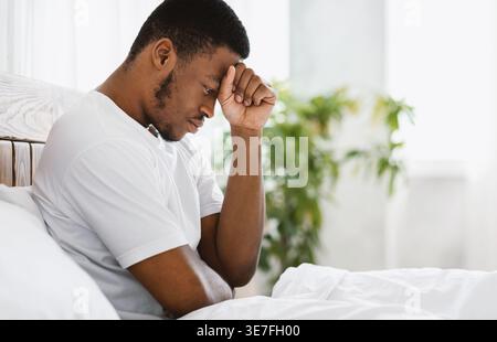 Uomo nero stressato in camicia bianca con la testa appoggiata sulla mano seduta sul letto di casa Foto Stock