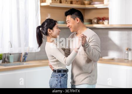 Allegra coppia asiatica che si tiene per mano danzando insieme in cucina a casa Foto Stock