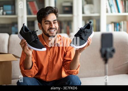 Vlogger maschile mediorientale che mostra i follower nuovissimi scarpe in streaming dal vivo seduti sul divano di casa Foto Stock