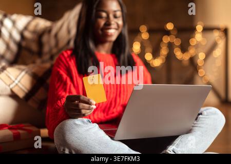 Adolescente nera che tiene in mano la carta di credito mentre utilizza un computer portatile seduto con le gambe incrociate sul pavimento in salotto con le luci natalizie a casa Foto Stock