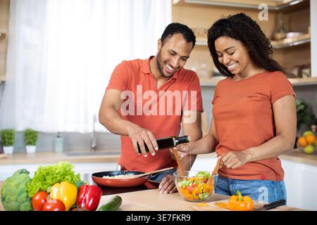 Coppia nera che versa l'olio per preparare l'insalata di verdure insieme mentre si trova al banco della cucina a casa Foto Stock