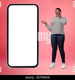 Donna nera con bocca aperta che tocca il viso che punta verso uno smartphone di grandi dimensioni mentre si trova sullo sfondo rosa in studio Foto Stock