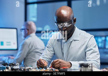 Ingegnere informatico maschio nero in occhiali protettivi e camice da laboratorio che lavora sulla scheda madre seduto al tavolo vicino al collega in laboratorio Foto Stock
